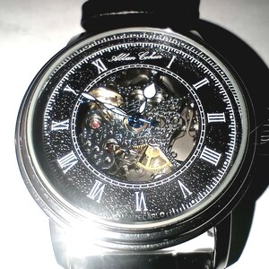 May include: A close-up of a silver and black Allan Cohen watch. The watch face has Roman numerals and a skeleton design, revealing the inner workings. The watch has a black leather band.