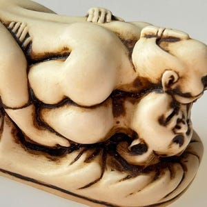 May include: A small, off-white carved figurine depicting two figures in an intimate embrace, resting on a textured, oval-shaped base. The sculpture features smooth, rounded forms with dark, recessed detailing that highlights the contours of the bodies.