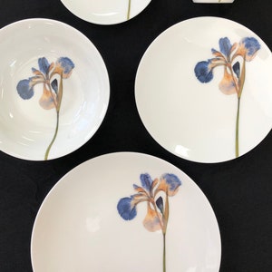 May include: A set of white porcelain dinnerware with a blue and orange iris flower design. The set includes a mug, a small plate, a large plate, a bowl, and a dinner plate.