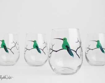 May include: Four clear glass wine glasses with a green hummingbird design on each. The hummingbirds are perched on a branch with black outlines.