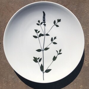 May include: A white ceramic plate with a pressed flower design. The flower is a dark green with a light purple bloom.