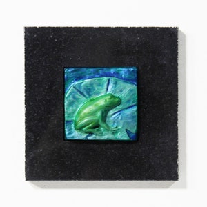May include: A green frog sculpture on a blue and green background, framed in black.