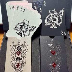 Pu&ograve; includere: Un mazzo di carte da gioco dal design unico. Le carte presentano un'illustrazione in bianco e nero su sfondo nero. Le facce delle carte sono bianche con semi e numeri neri. Gli astucci sono argentati e neri con un design rosso e nero.