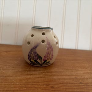 May include: A small, white ceramic vase with a floral design. The vase has a round shape and is decorated with purple and green flowers. It has small holes around the sides.