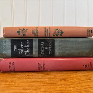 Puede incluir: Tres libros antiguos apilados sobre una superficie de madera. El libro superior es de color melocotón con detalles florales verdes y el título "The Little Locksmith". El libro del medio es gris con "The Silver Chalice". El libro inferior es rojo con "Little Women".