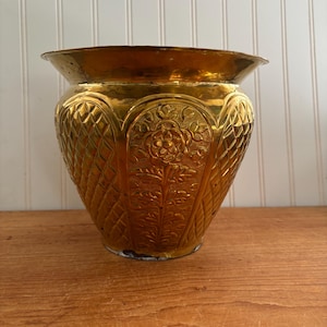 May include: A gold metal planter with an ornate design. The planter has a wide mouth and a rounded body. The design features a floral pattern and a geometric pattern.