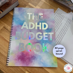 May include: A spiral-bound budget book with a watercolor-style cover in shades of pink, blue, and purple. The text THE ADHD BUDGET BOOK is printed in large, reflective silver letters. A circular white sticker notes 80 page spiral-bound.