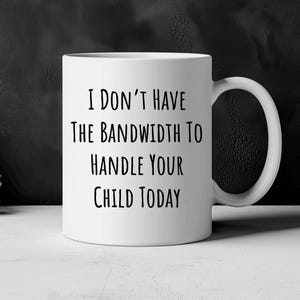 Puede incluir: Taza de cerámica blanca con asa negra. La taza tiene el texto "I DON'T HAVE THE BANDWIDTH TO HANDLE YOUR CHILD TODAY" en negro, con una fuente de estilo manuscrito. La taza está sobre un fondo oscuro.