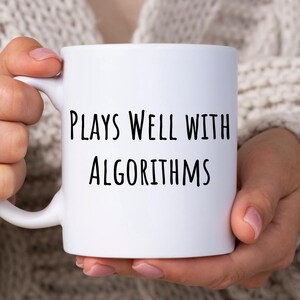 Op de afbeelding: Witte keramische koffiemok met zwarte tekst die "Plays Well With Algorithms" luidt.