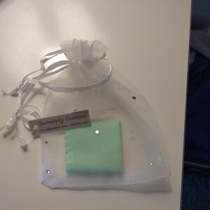 Puede incluir: Una bolsa de organza blanca con un pa&ntilde;o de limpieza verde en su interior. La bolsa tiene una peque&ntilde;a etiqueta blanca con el texto "Serendipity Creations" y una direcci&oacute;n de sitio web.