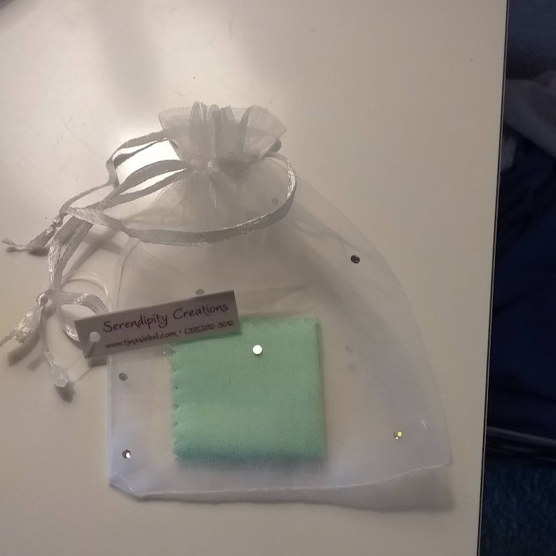 Puede incluir: Una bolsa de organza blanca con un pa&ntilde;o de limpieza verde en su interior. La bolsa tiene una peque&ntilde;a etiqueta blanca con el texto "Serendipity Creations" y una direcci&oacute;n de sitio web.