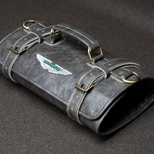 May include: A grey leather bag with a handle and straps, featuring a white and green emblem. The bag has brass-coloured hardware and white stitching details. The bag is rectangular and appears to be made of distressed leather.