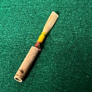 May include: A wooden clarinet reed with a red, yellow, and green wrapped section.