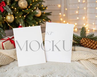 May include: Two blank 5x7 inch white cards with the word "MOCKUP" printed on them, resting on a cream-colored knitted blanket. A Christmas tree with ornaments and a gift box with a red ribbon are in the background. A pine cone and fairy lights add to the festive scene.