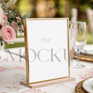 May include: A gold-framed sign with a white insert, measuring 12.7 x 17.8 cm, stands on a wooden base. The sign is set on a table with a floral tablecloth, near a vase of pink and white flowers and glassware.