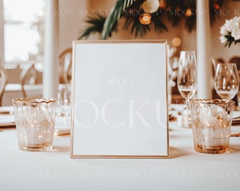 May include: A gold-framed blank sign sits on a white tablecloth with two clear glass candle holders on either side. The sign is on a table set for a wedding reception.