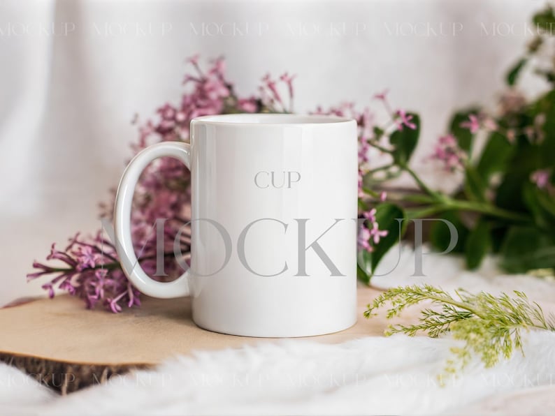 May include: A white ceramic mug with a curved handle, labeled "CUP" in gray text. The mug is surrounded by purple flowers and greenery, resting on a wooden slice and white fur. The overall aesthetic is clean and minimalist.
