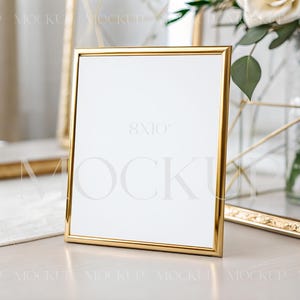 Puede incluir: Un marco de fotos dorado con interior blanco, ideal para exhibir fotos o obras de arte. El marco es rectangular y se apoya sobre una superficie de color claro. En el fondo se ve un vaso con un diseño decorativo.