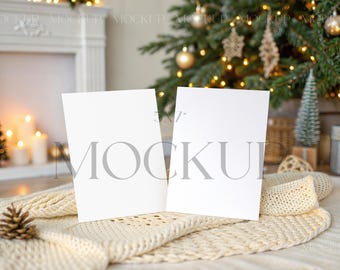 May include: Two blank white cards, likely for a greeting or invitation, are propped up on a cream-colored knitted blanket. A pine cone and a small Christmas tree are in the foreground. A decorated Christmas tree and fireplace are in the background.