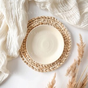 May include: A white ceramic plate on a woven straw placemat with a white cloth and dried pampas grass on a white background.