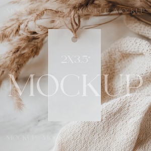 May include: A white gift tag with a hole punched in the top and the text "2x3.5" printed on it. The tag is lying on a white marble surface with a beige knitted fabric and dried pampas grass.