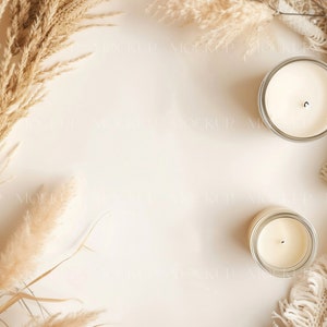 May include: Two white candles in glass jars on a beige background with dried pampas grass and a white crocheted throw.