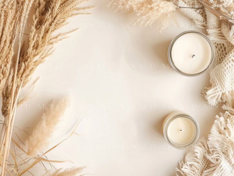 May include: Two white candles in glass jars on a beige background with dried pampas grass and a white crocheted throw.