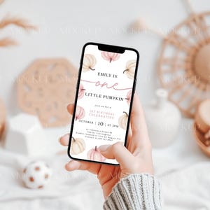 May include: A mobile phone displaying a digital invitation for a first birthday party. The invitation features a watercolor illustration of pumpkins and the text "Emily is one little pumpkin".