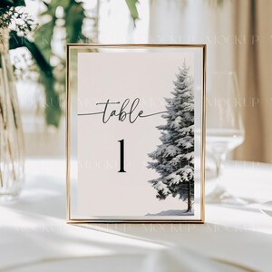 May include: A gold-framed table number card with a white background and black text. The card says "table 1" and features a watercolor illustration of a snow-covered pine tree.