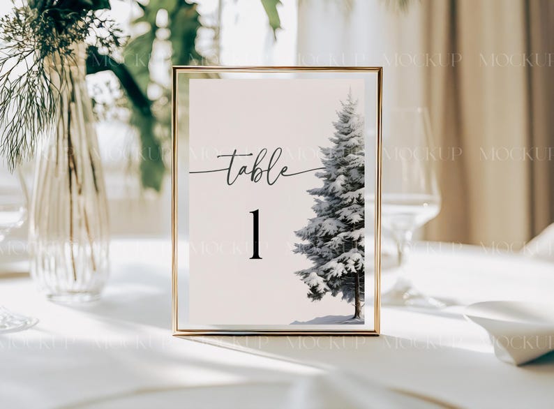 May include: A gold-framed table number card with a white background and black text. The card says "table 1" and features a watercolor illustration of a snow-covered pine tree.