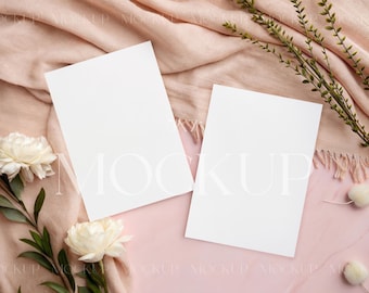 May include: Two blank white cards on a pink surface with a blush pink fabric and white flowers.