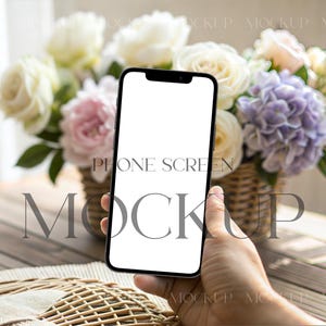 May include: A black smartphone with a blank white screen is held in front of a floral arrangement. The flowers include white roses, pink roses, and purple hydrangeas in a wicker basket. The words "PHONE SCREEN MOCKUP" are overlaid on the image.