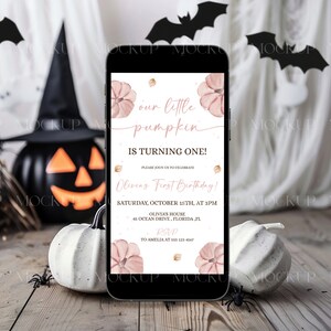 K&ouml;nnte beinhalten: Ein Smartphone zeigt eine Einladung zum Thema Halloween mit dem Text "our little pumpkin is turning one!" in rosa Schrift. Die Einladung zeigt Aquarellk&uuml;rbisse und goldene Akzente. Das Telefon liegt auf einer Holzoberfl&auml;che mit dekorativen K&uuml;rbissen und Spinnen.
