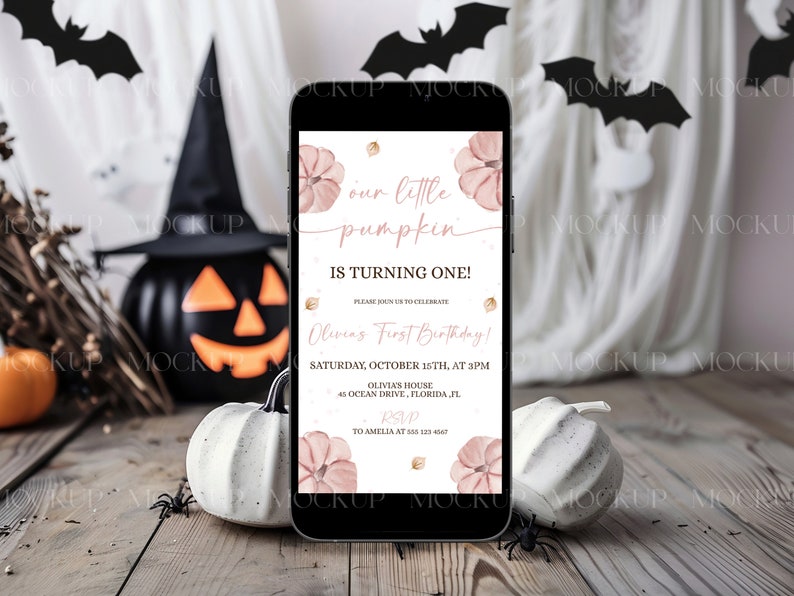 K&ouml;nnte beinhalten: Ein Smartphone zeigt eine Einladung zum Thema Halloween mit dem Text "our little pumpkin is turning one!" in rosa Schrift. Die Einladung zeigt Aquarellk&uuml;rbisse und goldene Akzente. Das Telefon liegt auf einer Holzoberfl&auml;che mit dekorativen K&uuml;rbissen und Spinnen.