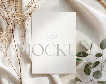 Puede incluir: Maqueta de tarjeta blanca de 5x7 pulgadas con la palabra "MOCKUP" en gris claro. La tarjeta está sobre una superficie de tela blanca, con flores secas y hojas verdes a ambos lados. La iluminación suave crea sombras.