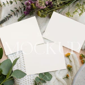 Puede incluir: Tres tarjetas en blanco de 12,7 x 8,9 cm dispuestas sobre una manta blanca de punto de cable, rodeadas de flores frescas y vegetación. El arreglo incluye rosas naranjas, flores silvestres moradas y ramitas de eucalipto.