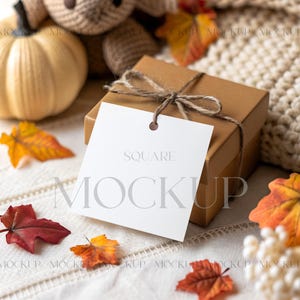 May include: A brown square gift box tied with twine, featuring a white square tag with the word "SQUARE MOCKUP" printed on it. The scene includes a decorative pumpkin, fall leaves, and a knitted blanket.