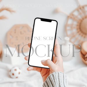 May include: A hand holding a black smartphone with a blank white screen. The phone is in a neutral background with a blurred image of a wicker basket and other decor.