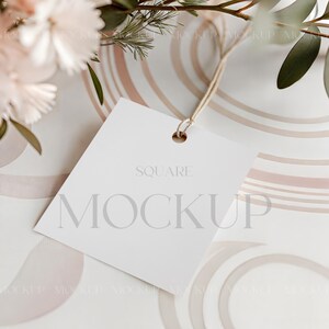 Pode incluir: Uma etiqueta quadrada branca com as palavras "SQUARE" e "MOCKUP" impressas, pendurada por um fio bege. A etiqueta está sobre uma superfície estampada com detalhes florais e de folhas.