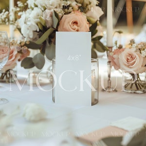 May include: A white card with the text "4x8" printed on it is displayed on a table setting. The card is surrounded by pink and white flowers in glass vases.