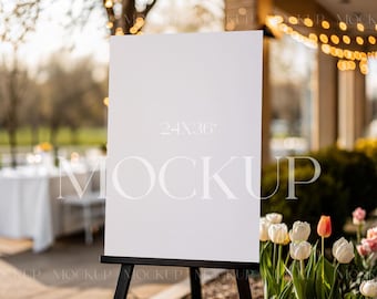 May include: A white sign on an easel with the text "24x36" and "MOCKUP" in a light gray font. The sign is in front of a blurred background of a patio with white tables and chairs, green plants, and string lights.