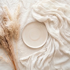 May include: A white ceramic plate sits on a white fabric background with a bouquet of dried pampas grass to the left.