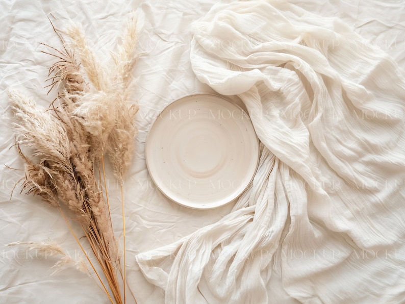 May include: A white ceramic plate sits on a white fabric background with a bouquet of dried pampas grass to the left.