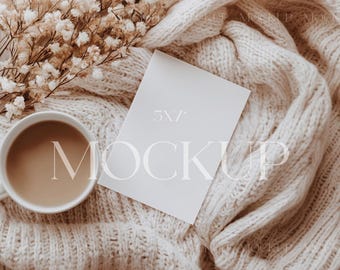 May include: A white card mockup with the text "5x7" and "MOCKUP" on a cream knitted blanket with a cup of coffee and dried flowers.