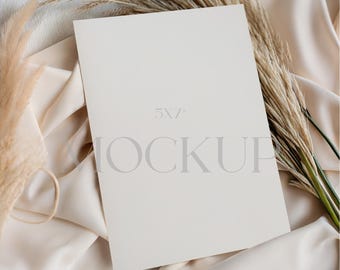 Puede incluir: Una maqueta de tarjeta en blanco de 5x7 pulgadas con la palabra "MOCKUP" impresa en ella, sobre una tela de satén color crema. Hierba de la pampa seca y otros elementos decorativos están dispuestos alrededor de la tarjeta, creando una estética suave y neutra.
