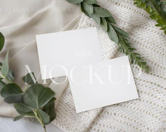 May include: Two blank white 5x3.5 inch cards are arranged on a cream-colored knitted blanket and draped fabric. Green leafy sprigs are placed around the cards. The word "MOCKUP" is faintly visible on the cards.