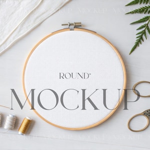 Könnte beinhalten: Ein runder Stickrahmen mit weißem Stoff, auf dem die Wörter "ROUND" und "MOCKUP" aufgedruckt sind. Goldene Scheren, Garnrollen und ein Farnzweig sind um den Reifen herum angeordnet. Ein weißes Tuch befindet sich in der oberen linken Ecke.