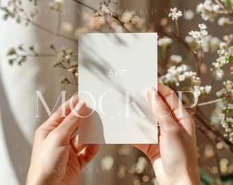 Può includere: Una persona tiene in mano un cartoncino bianco con la scritta "5x7" stampata su di esso. Il cartoncino è tenuto davanti a uno sfondo sfocato di fiori bianchi.