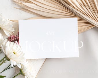 May include: A white card mockup with the text "MOCKUP" in white on a white background. The card is surrounded by dried palm leaves and white flowers.