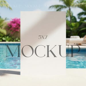 Op de afbeelding: Een witte 5x7 mockup kaart staat bij een zwembad met helder blauw water. Het woord "MOCKUP" is in een donkergrijze lettertype over de onderkant van de kaart gedrukt. Palmbomen en roze bloemen zijn zichtbaar op de achtergrond.